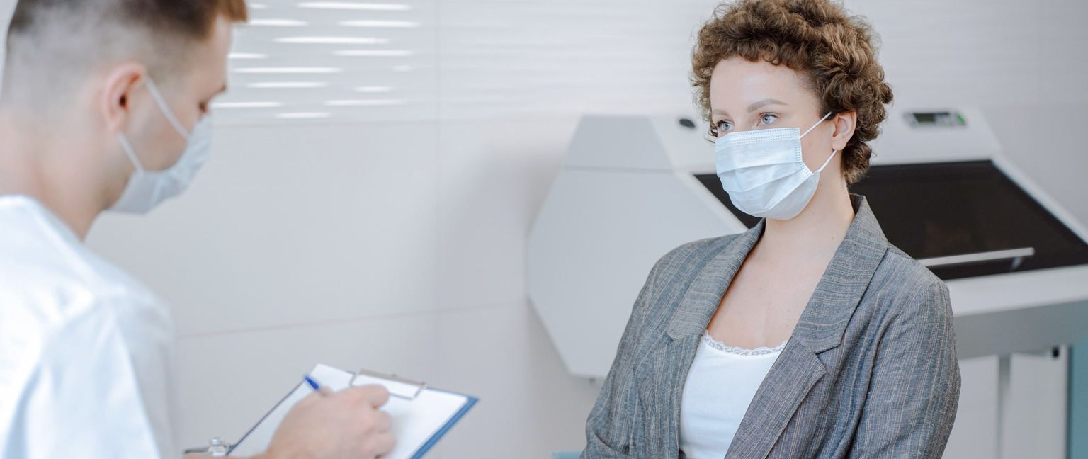 A woman in a mask sits talking to a masked doctor who is writing on a clipboard.