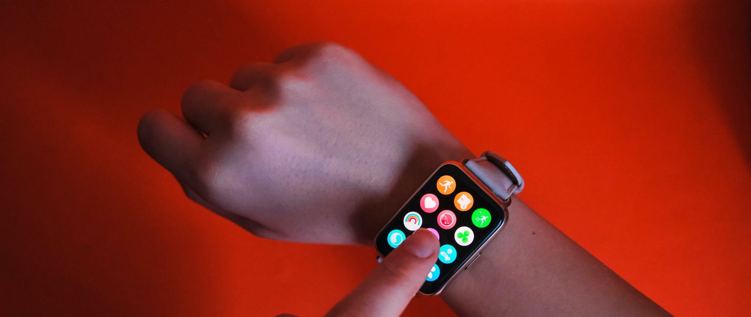 A finger is shown tapping the screen of an Apple Watch, which displays a grid of colorful icons. The hand is on a rust-colored background.