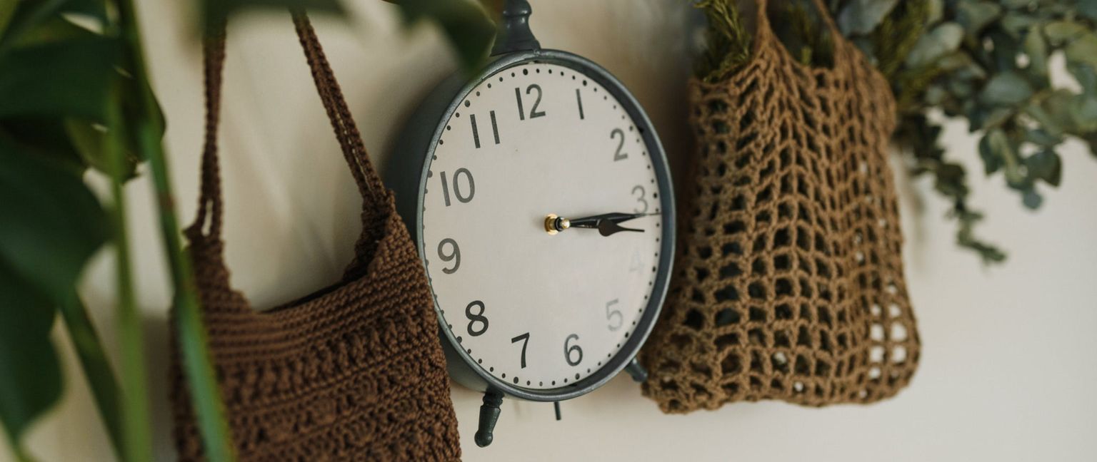 A vintage-style clock hangs on a wall between two crocheted bags. One bag is dark brown and solid, the other is light brown and a net bag.