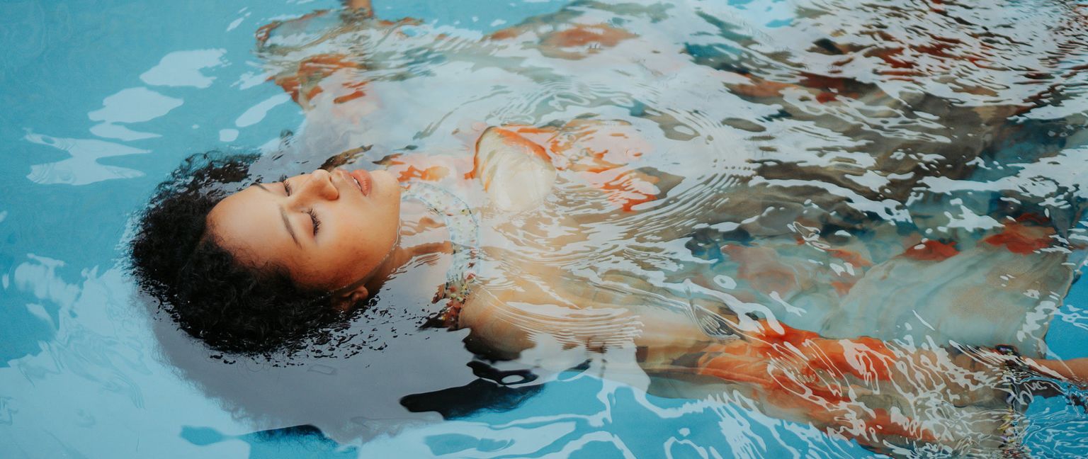 A woman with eyes closed floats on her back in clear blue water wearing a light colored dress with orange and blue floral print.