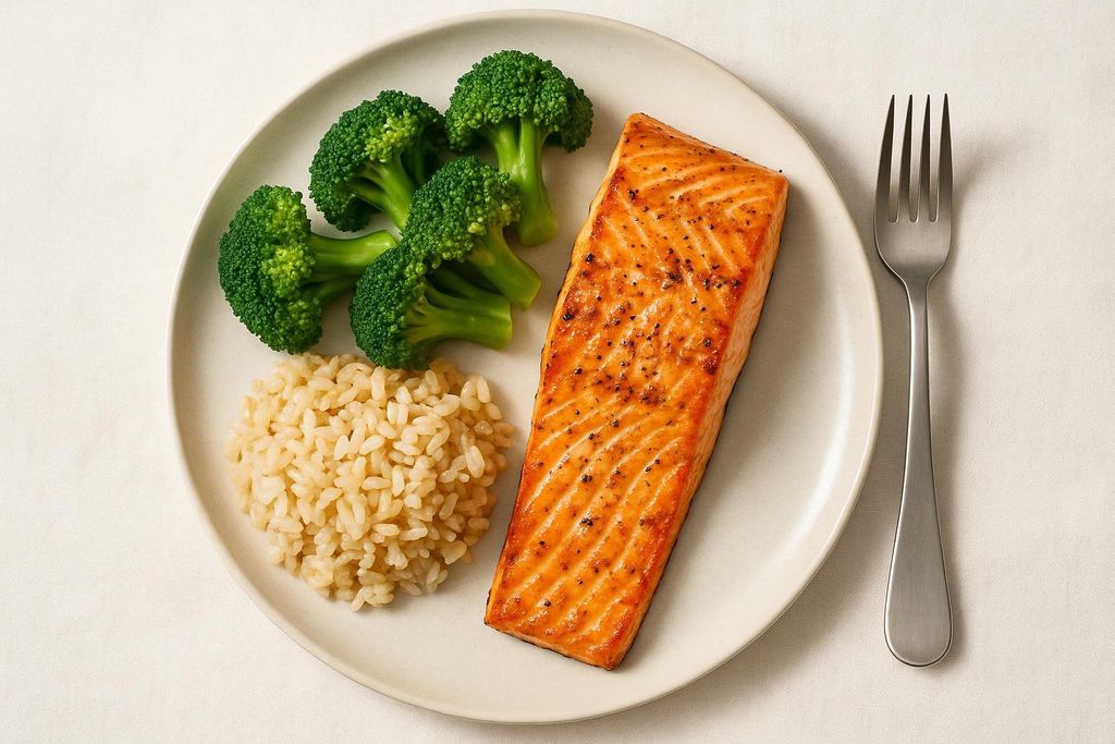 A balanced meal served on a light plate, featuring a seasoned grilled salmon fillet, a serving of steamed broccoli florets, and a portion of fluffy brown rice. A silver fork rests to the right of the plate.
