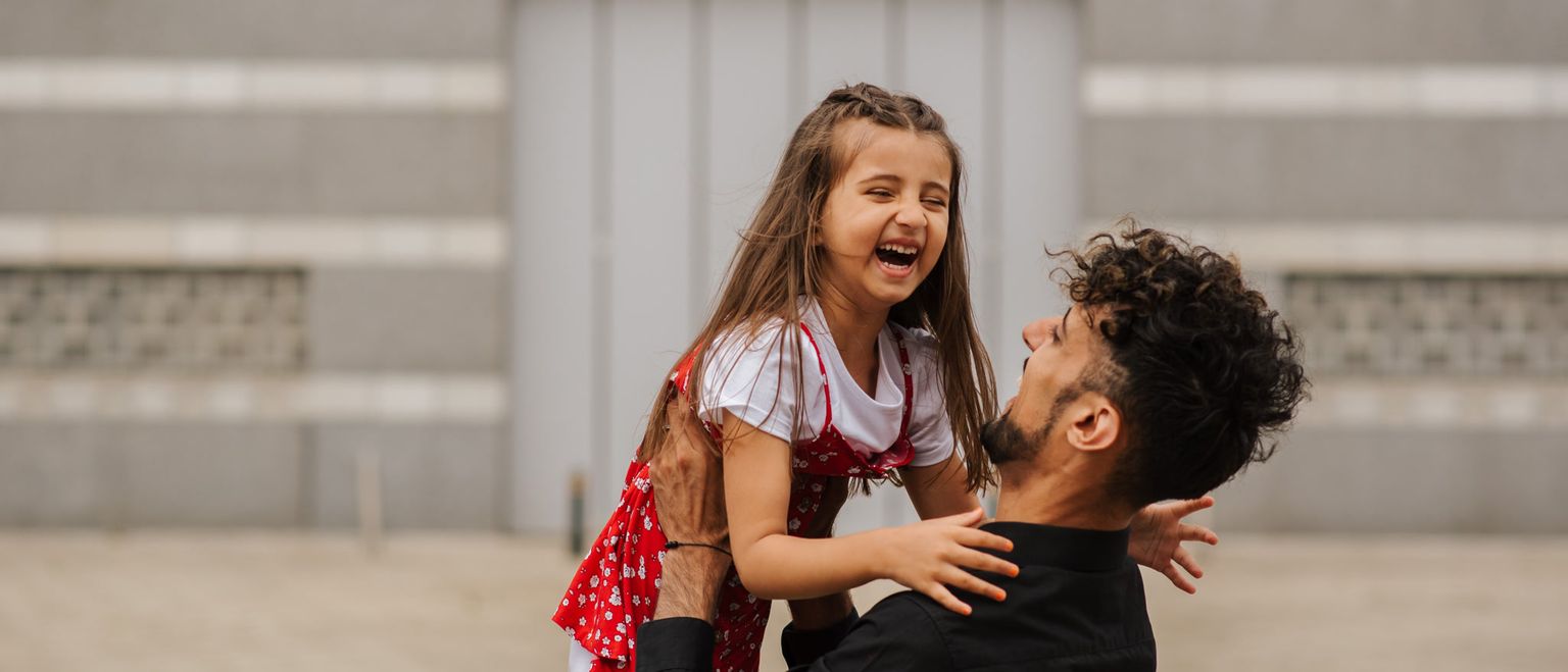 A father holds his young daughter up in the air. Both are laughing with their mouths wide open. The daughter is wearing a red and white dress and the father is wearing a black shirt.