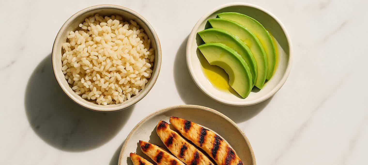 Overhead shot of a healthy meal including a bowl of brown rice, a bowl of sliced avocado drizzled with olive oil, and a plate of grilled chicken breast strips, all arranged on a white marble surface.