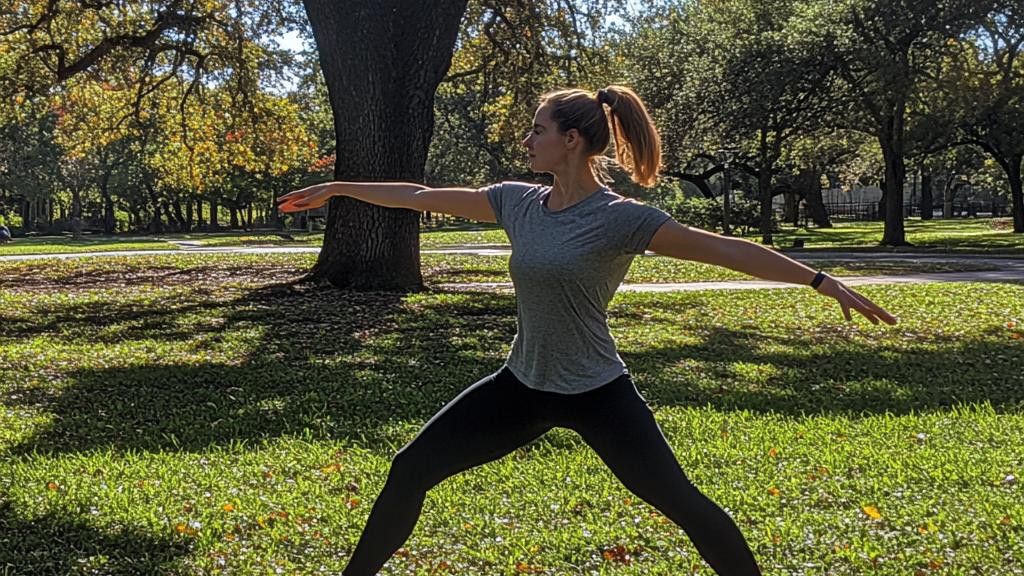 A woman with a ponytail demonstrates a warrior pose in an outdoor park. She is wearing a gray t-shirt and black leggings.