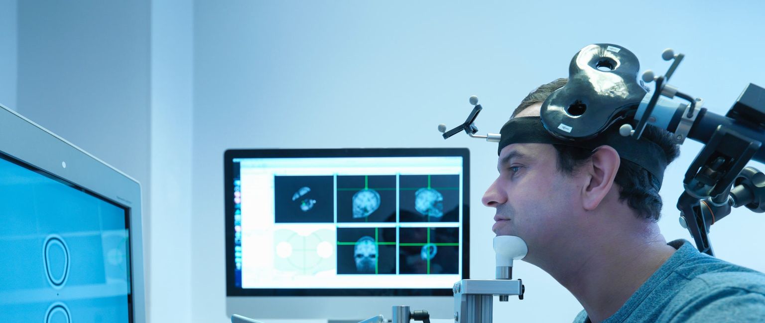 A man wearing equipment for transcranial direct stimulation, with a monitor showing brain scans and targeting information in the background.