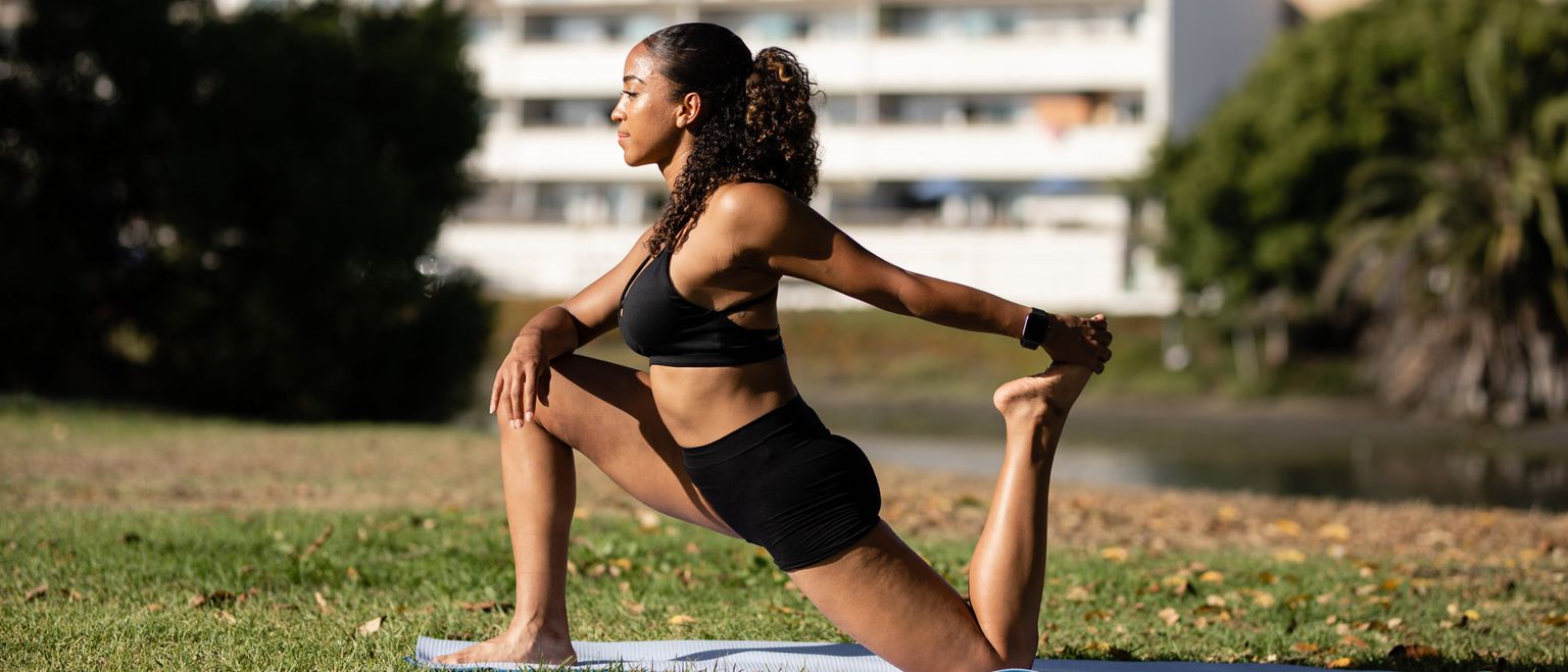 A fit woman in black workout clothes stretches on a yoga mat in a park. She is holding one foot behind her, stretching her quad muscle.