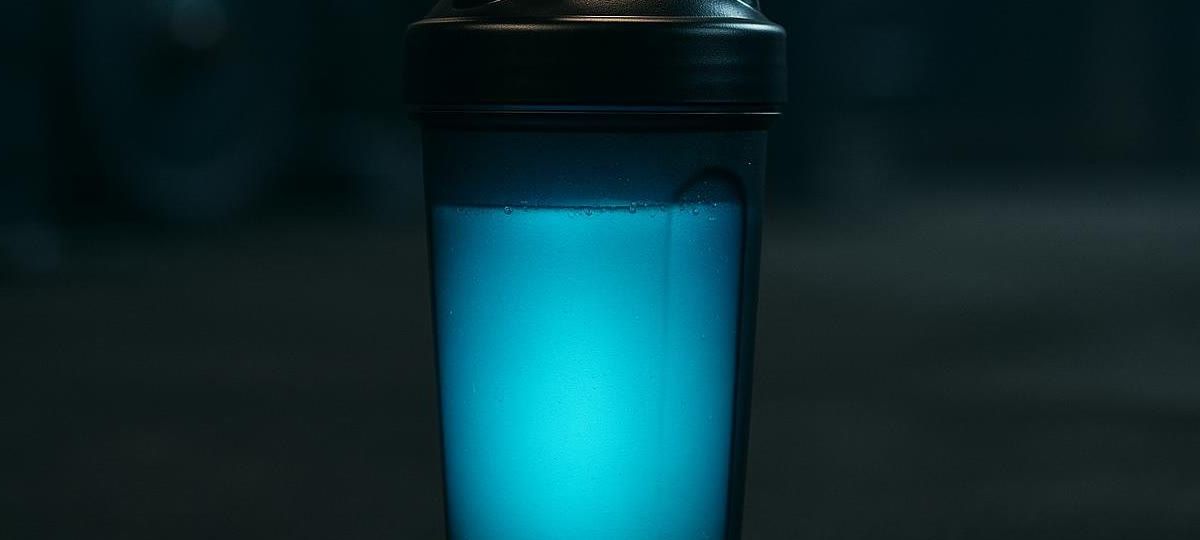 A close-up shot of a black shaker bottle filled with a bright blue, luminous pre-workout drink, sitting on a dark surface in what appears to be a gym.