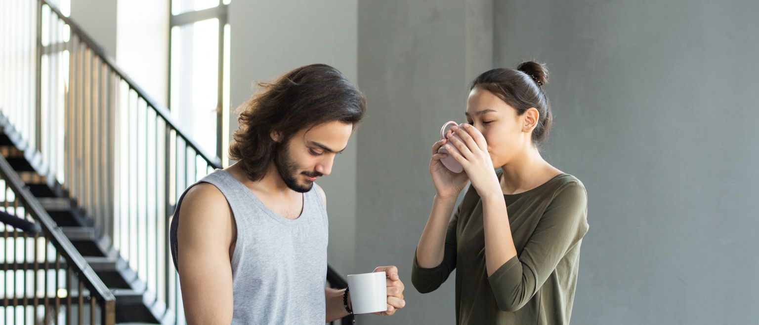 A man in a grey tank top holds a white mug and looks down, while a woman in a green shirt sips from a pink mug next to him.