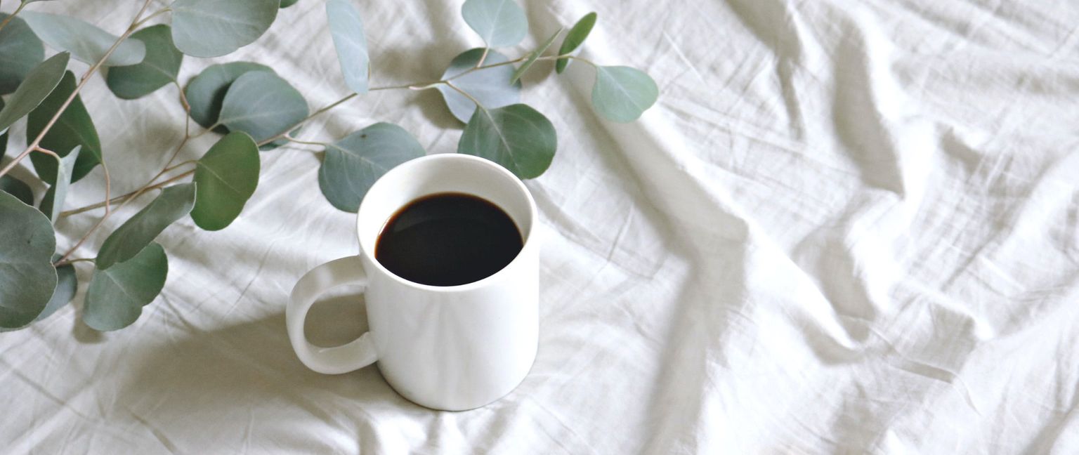 A white coffee mug sits on wrinkled white sheets. A branch of eucalyptus leaves is to the left of the mug.