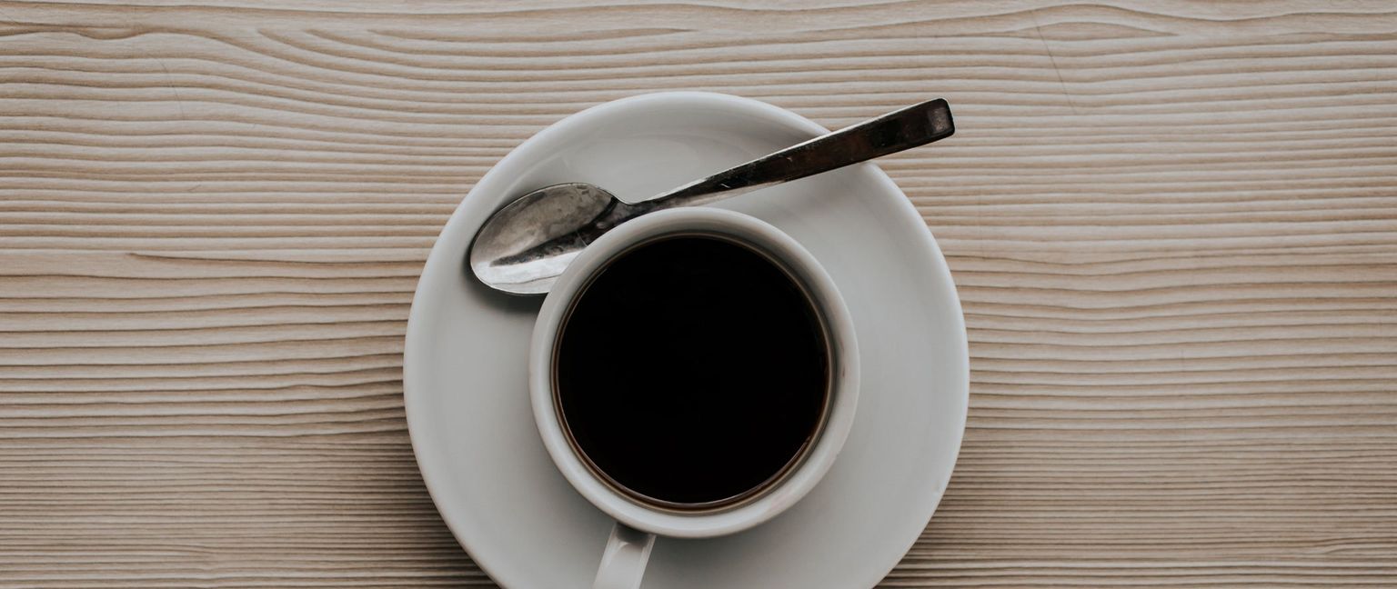 A top-down view of a cup of black coffee and a spoon sitting on a white saucer, on a light-colored wood grain table.