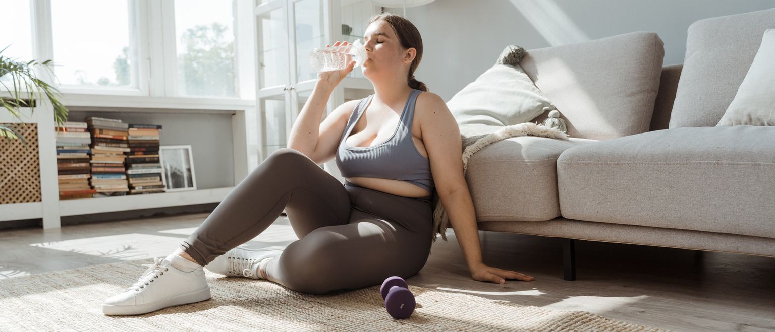 A woman in workout clothes sits on the floor after exercising, drinking from a glass of water.