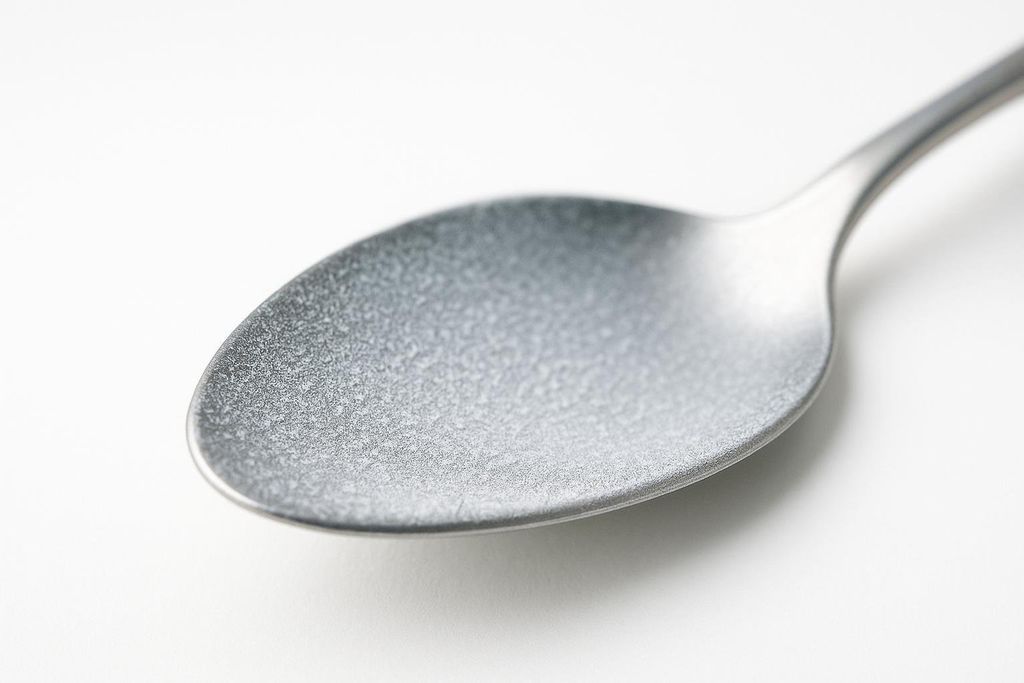 A close-up shot of the bowl of a frosted metal spoon on a white background. The spoon's surface appears textured and matte.