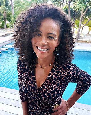 A woman with curly hair smiles at the camera with a pool behind her.