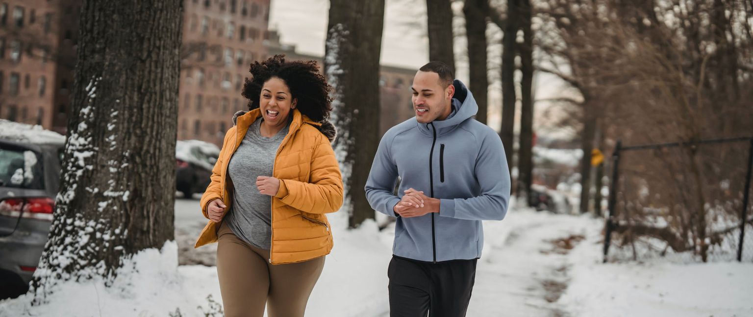 Two people are smiling and running on a snowy sidewalk. The woman on the left wears a bright yellow coat, and the man on the right wears a blue hoodie.