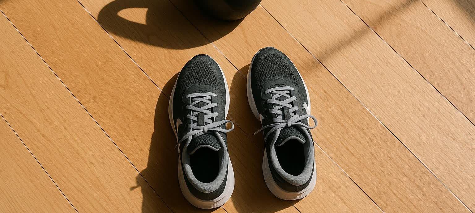 A pair of grey running shoes with white soles and grey laces resting on a light wooden floor. A black kettlebell is partially visible in the upper left corner, casting a shadow.