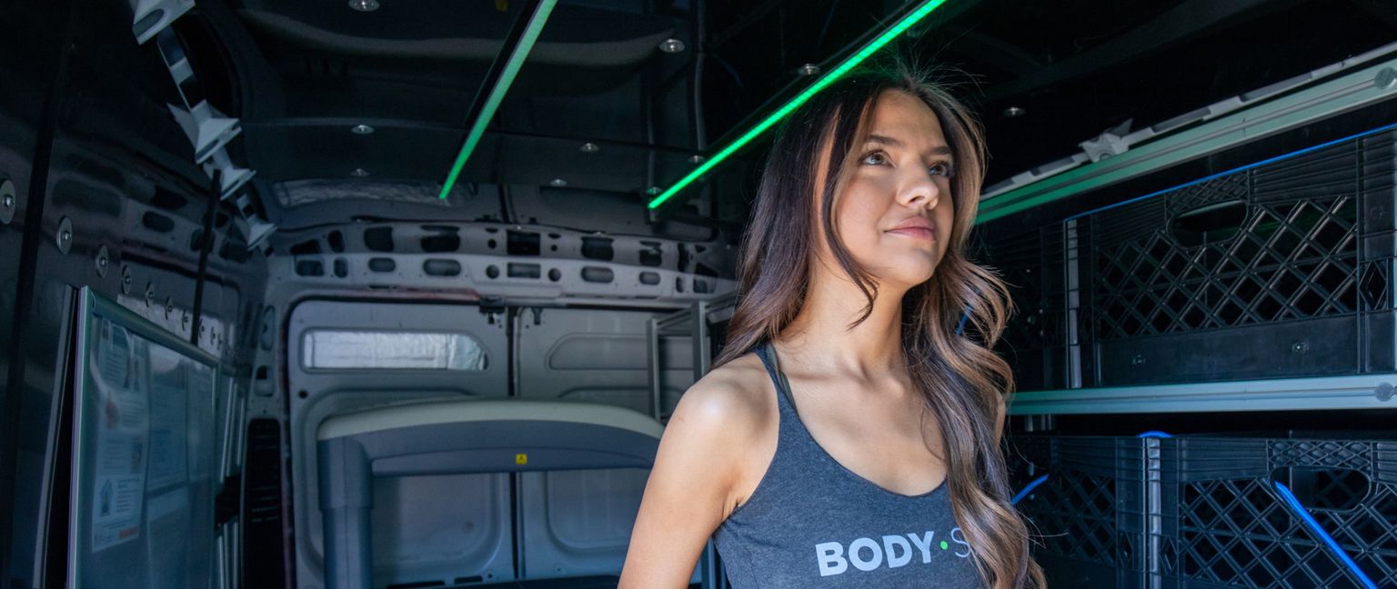 A young woman in a gray tank top stands inside the back of a mobile clinic van looking ahead.