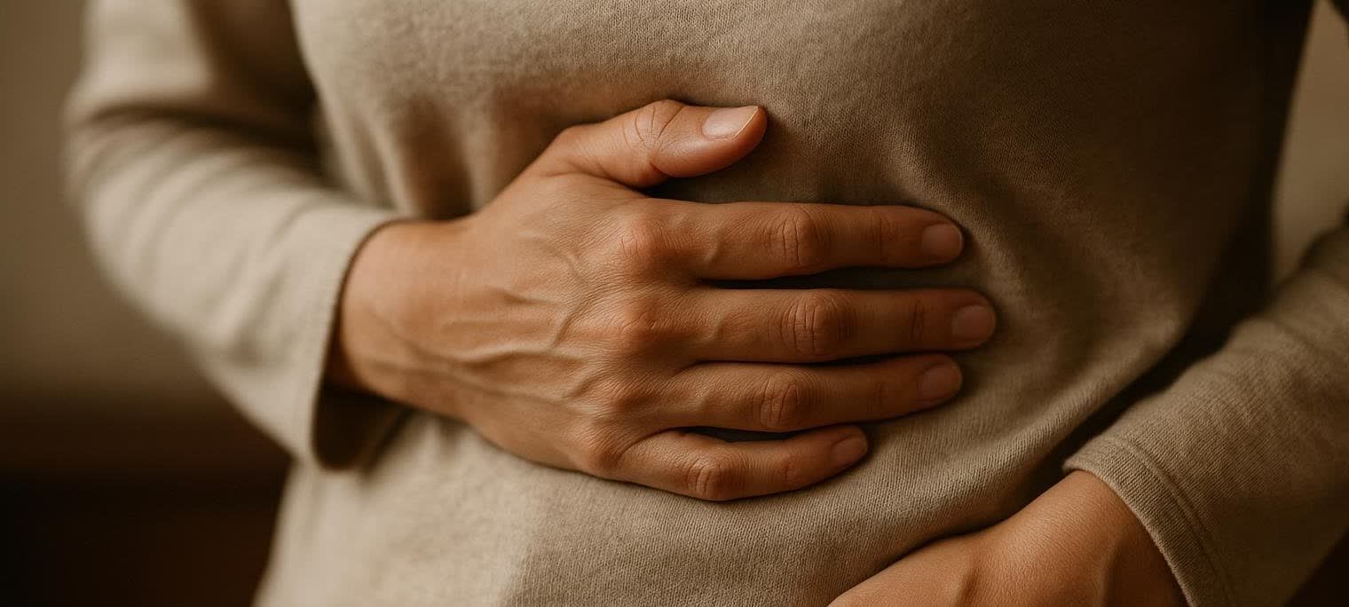 Close-up of a person wearing a beige long-sleeved shirt, holding their stomach with a hand, indicating discomfort or pain. The hand has visible veins.