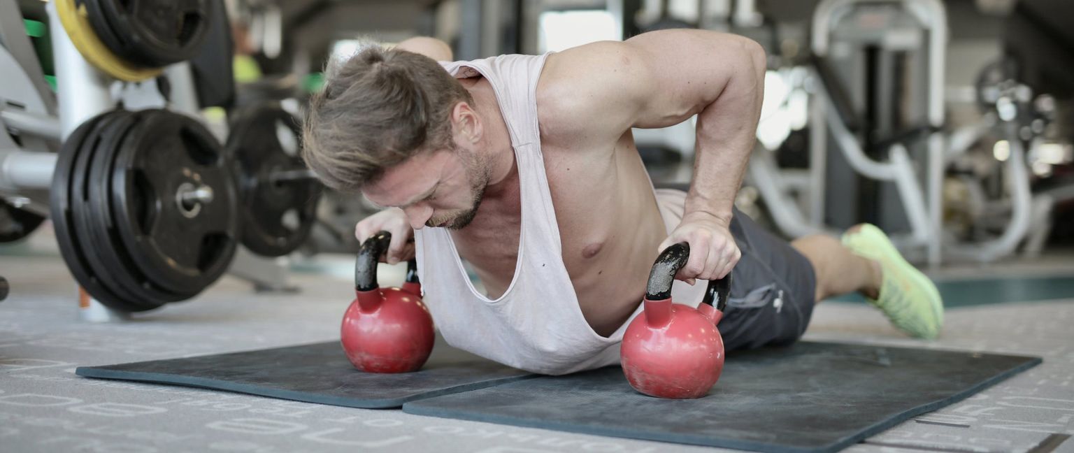 A fit man in a white tank top and shorts is doing push-ups on two red kettlebells in a gym.