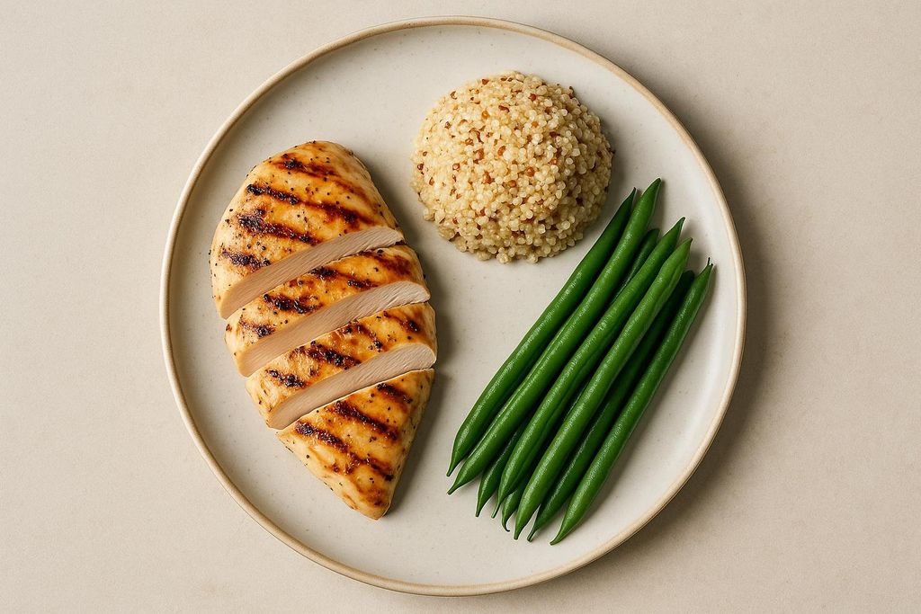 A plate of grilled chicken breast, sliced, served with a side of quinoa and a pile of green beans. The chicken has distinct grill marks.