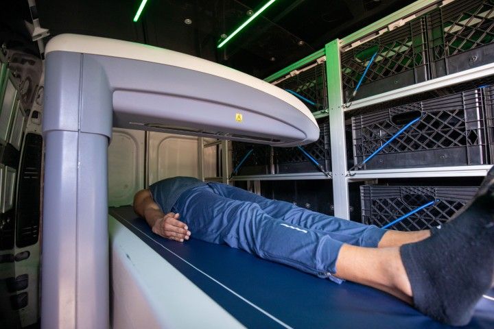 A person lies face down on a BodySpec DEXA scanner inside a mobile van. The scanner is a large, light grey and white machine, and the person is wearing a dark blue shirt and pants. The interior of the van has shelves with black crates.