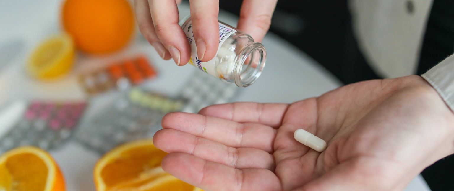Closeup of a hand holding a single white pill. In the background, a bottle of pills, oranges, and blister packs of pills are visible.