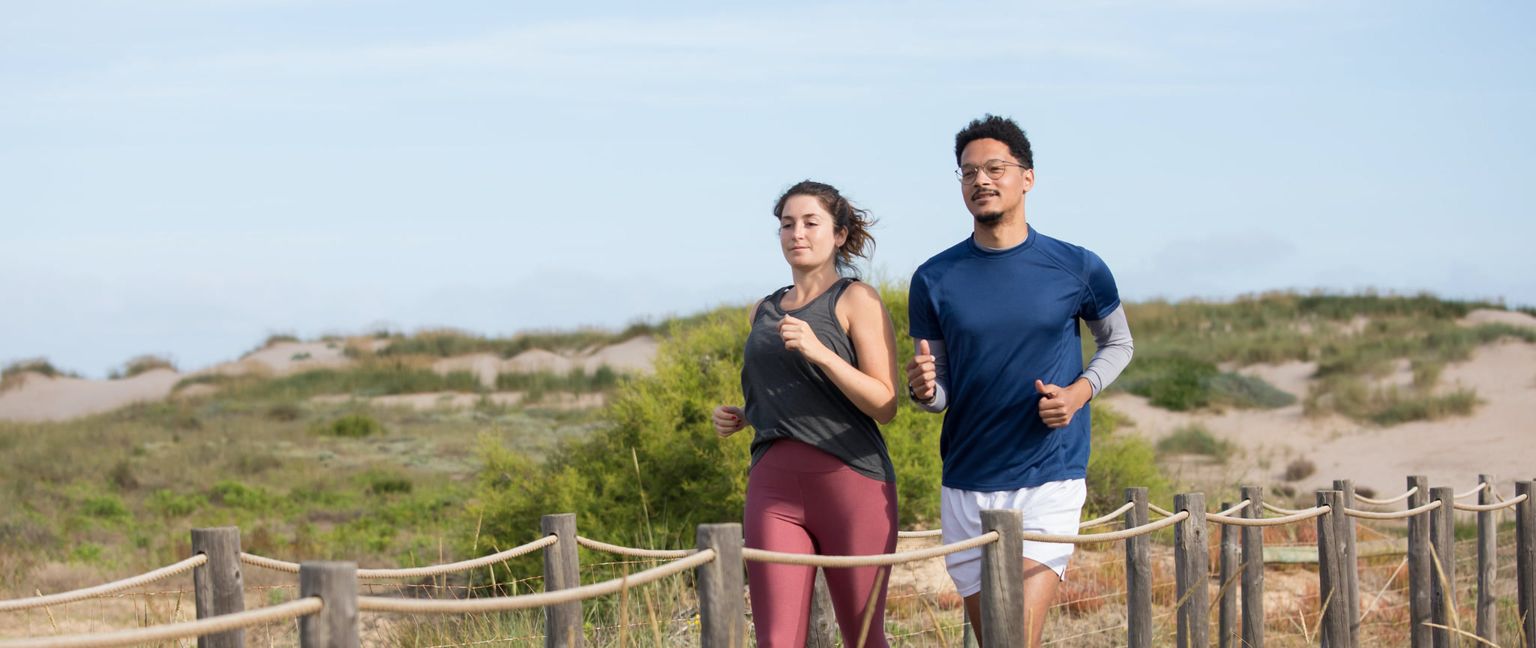 A man and woman jog together on a dirt path, passing by dunes.