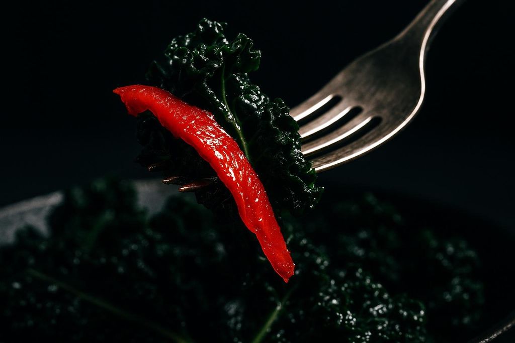 Close-up of a fork spearing a piece of deep red pepper and dark green kale, set against a dark, blurred background of more kale.