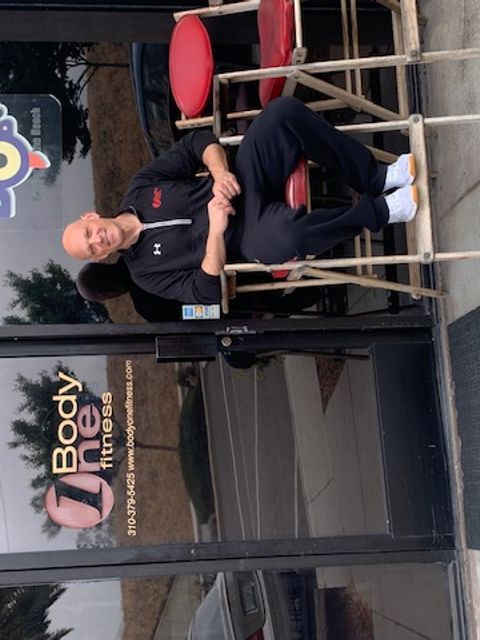 Lou sitting on a slanted metal chair outside of a fitness gym called Body One Fitness.