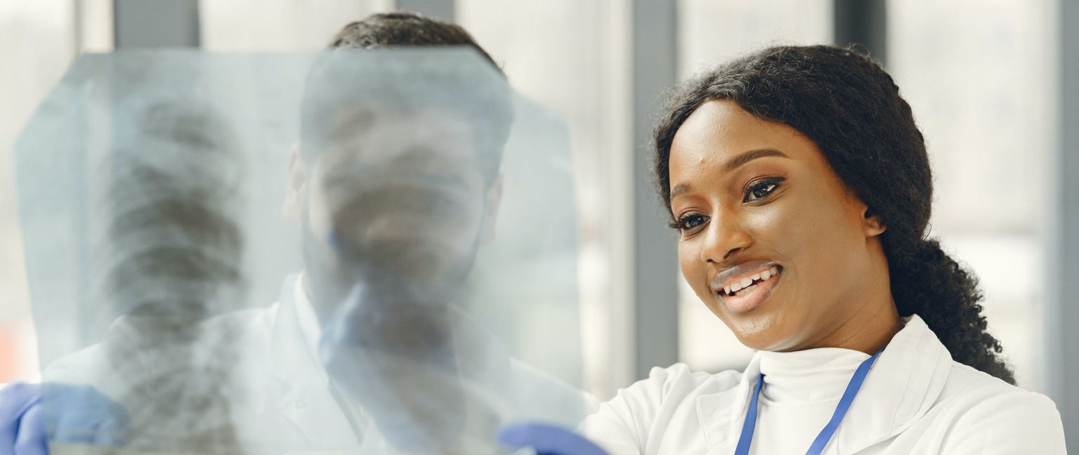 Two doctors, one in focus smiling, examine a large illuminated X-ray.