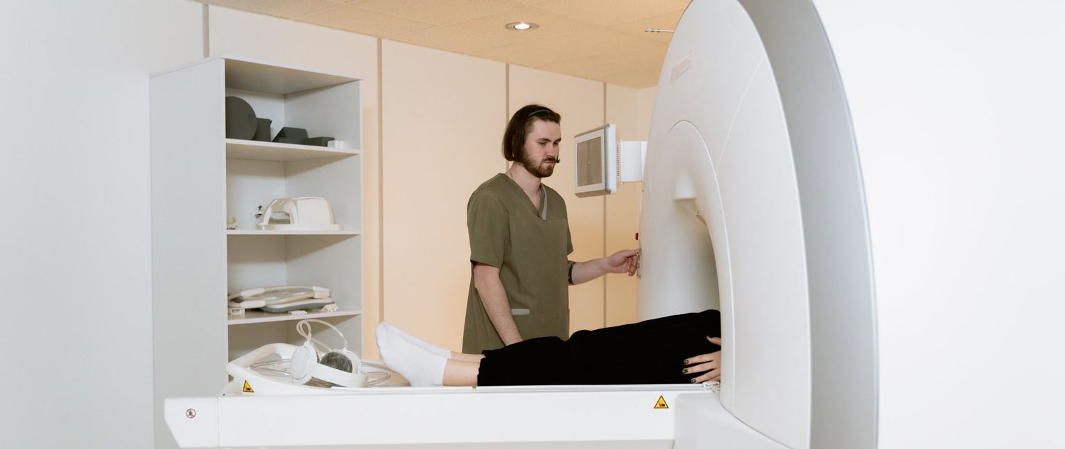 A medical technician wearing a green scrub shirt is helping a patient on the bed of a CT scanner. The patient is lying face-up with their lower body visible, covered by a black blanket and white socks. The technician is standing next to the machine, adjusting controls on a small screen.