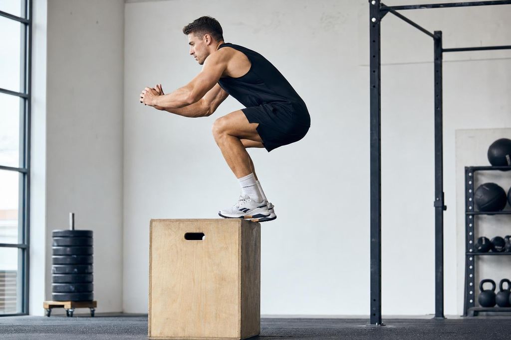 A male athlete in black workout clothes is performing a plyometric box jump, mid-air onto a wooden box. He is in a modern gym with a stack of weights and other fitness equipment in the background.