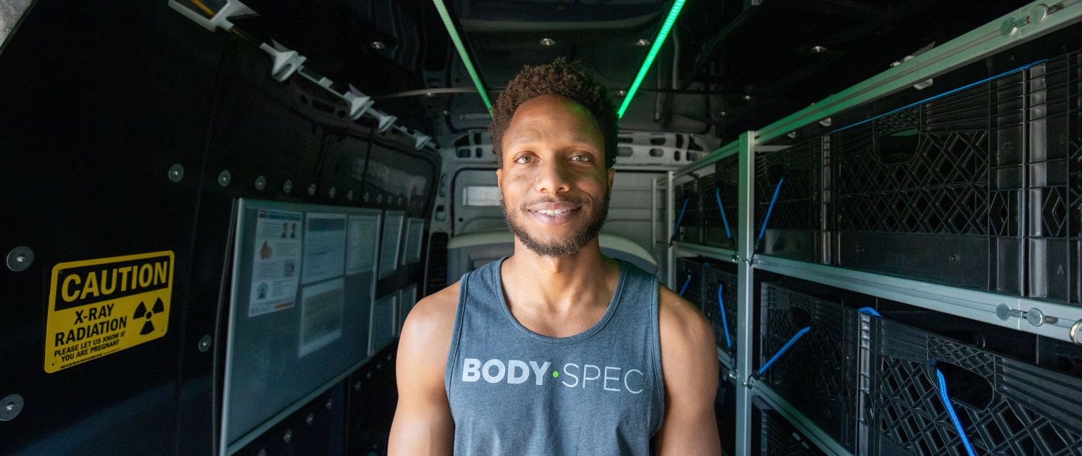 A man wearing a Body Spec tank top stands inside a DEXA scanning van. A caution sign for x-ray radiation is visible on the wall behind him.