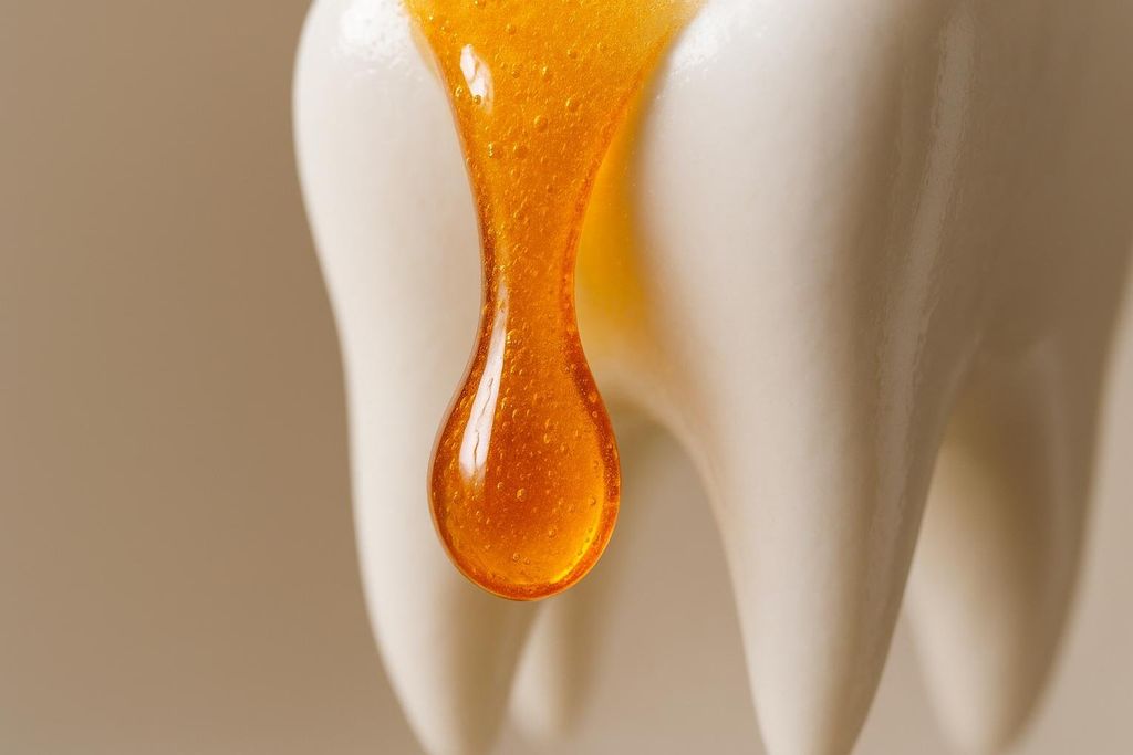 An abstract macro photo showing a sticky, acidic-looking liquid, resembling honey or syrup, dripping down a smooth white surface shaped like a tooth, symbolizing the effect of sugary or acidic substances on tooth enamel.