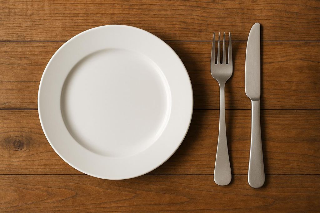 A pristine white plate, fork, and knife are neatly arranged on a warm, dark wooden table, viewed from directly above. The plate is empty, and the cutlery is made of brushed metal.